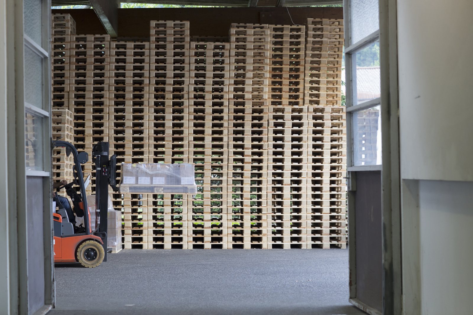 An orange forklift carrying a wrapped, labelled load in the doorway of a storage building, with tall stacks of timber pallets behind it