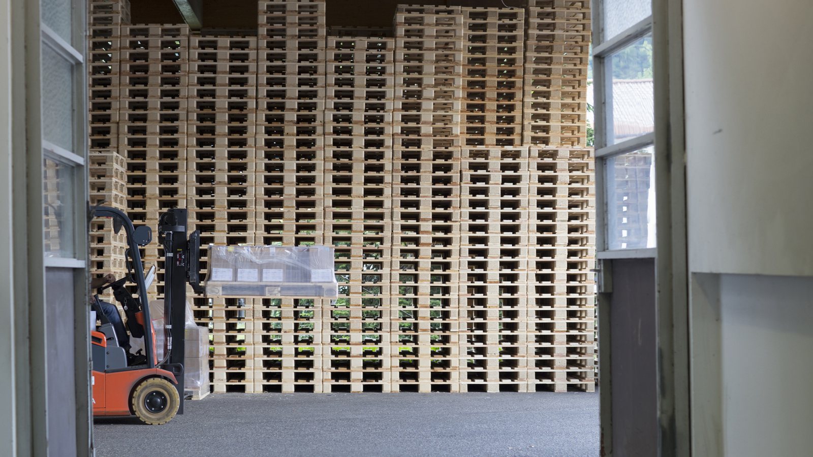 A forklift carrying a wrapped load past tall stacks of timber pallets in a storage building